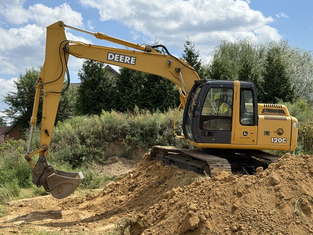 A John Deere excavator positioned on top of a dirt pile, with the operator in the cab, surrounded by trees and blue sky.