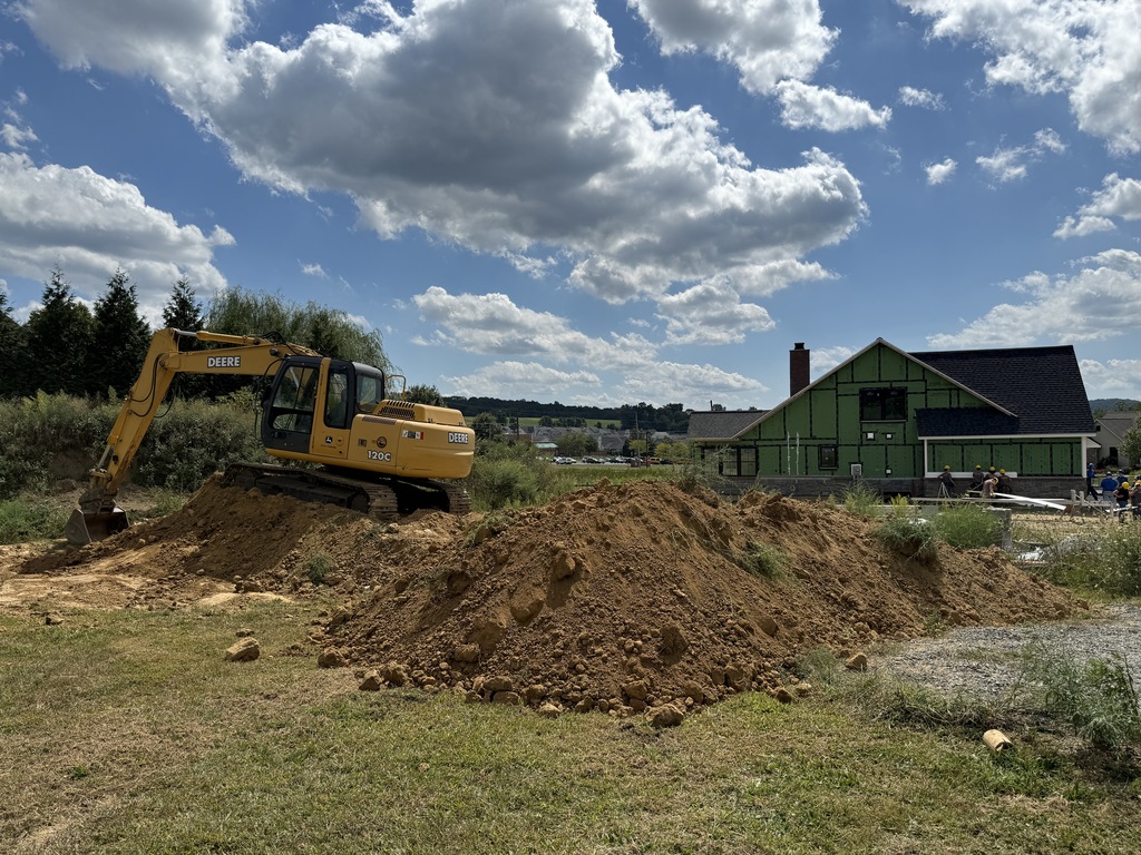 A wide shot of a John Deere excavator moving dirt piles in the foreground, with BCTC’s student-built house under construction and students working near the foundation in the background.