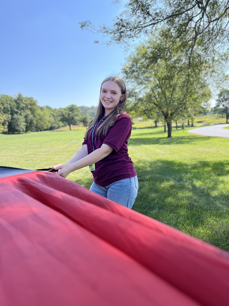 A student wearing a maroon polo shirt smiles while holding the edge of a bright red parachute on a sunny day, standing on green grass with trees in the background.