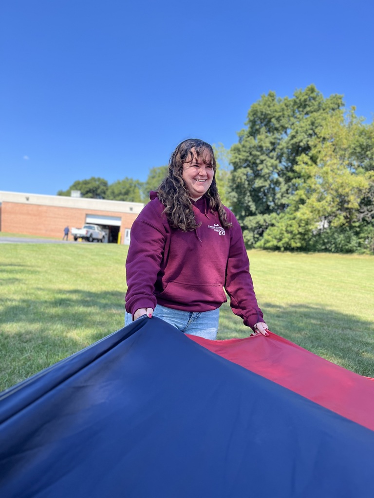 A student in a maroon BCTC hoodie holds onto the navy and red sections of a parachute, smiling outdoors with a school building and blue sky behind her.