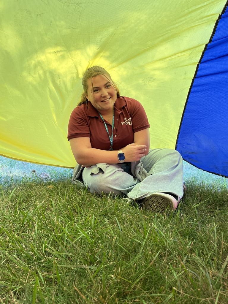 A student in a maroon polo shirt sits on the grass under the parachute’s yellow and blue sections, smiling and leaning slightly to the side.