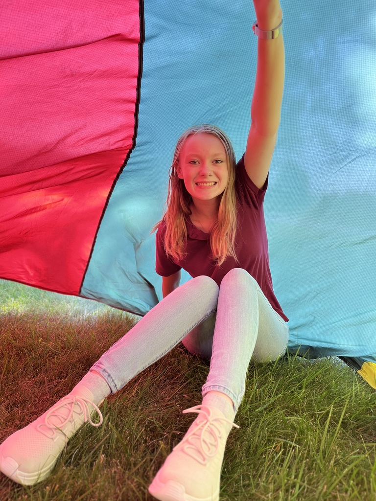 A student in a maroon polo shirt sits under the parachute with one arm raised, smiling as the red and blue fabric drapes overhead, with her legs stretched out on the grass.