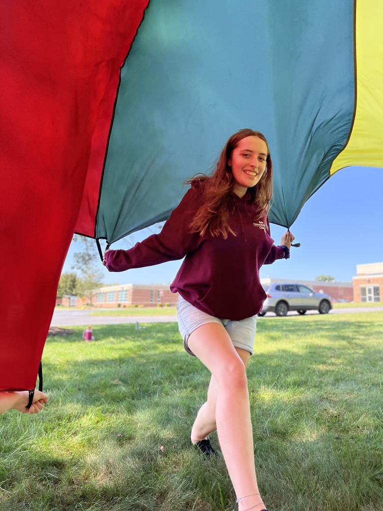 A student in a maroon hoodie runs under a colorful parachute, smiling as the red, blue, and yellow fabric arches overhead.