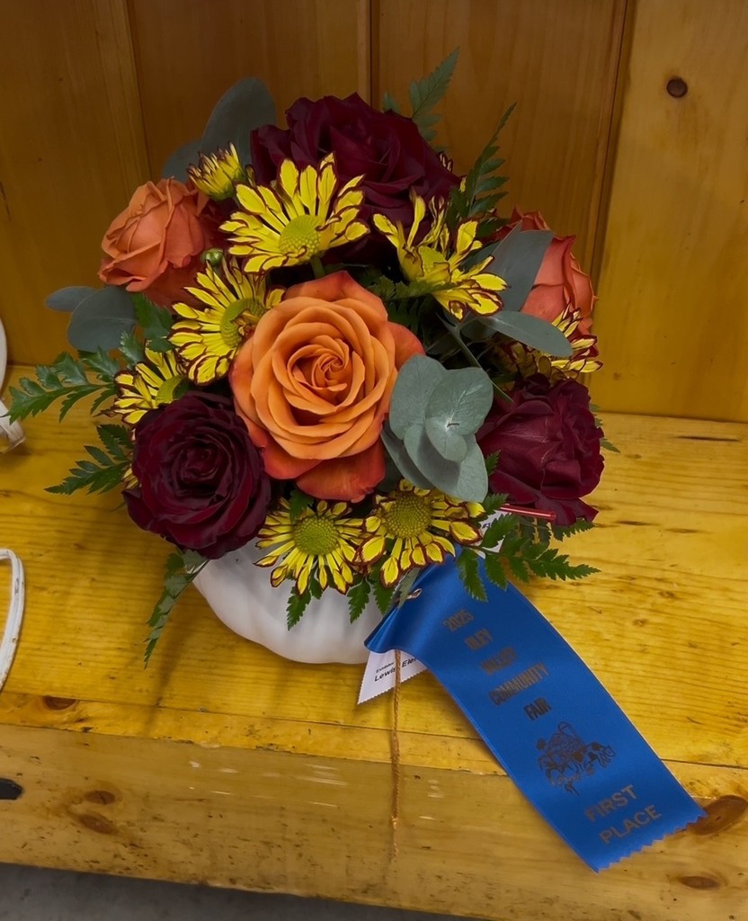 A compact bouquet in a white bowl-shaped vase with orange roses, red roses, yellow daisies, and greenery. A blue first-place ribbon from the 2025 Oley Valley Community Fair is attached with exhibitor tag reading “Elizabeth Lewis.”