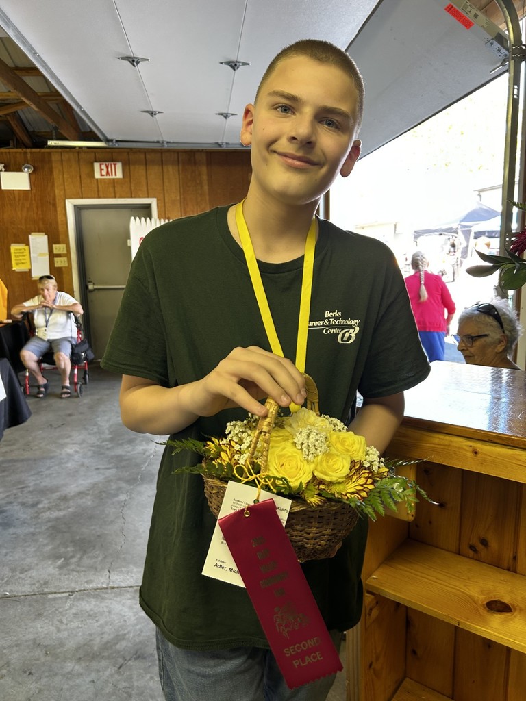 A young man, Michael Adler, holds a small basket arrangement with yellow roses and white baby’s breath. A red second-place ribbon from the 2025 Oley Valley Community Fair is attached.
