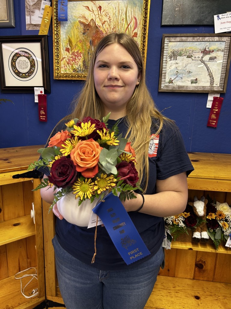 A young woman, Elizabeth Lewis, holds her floral arrangement of orange and red roses with yellow daisies in a white vase. A blue first-place ribbon is attached to the arrangement. She is standing in front of artwork and other floral entries at the fair.