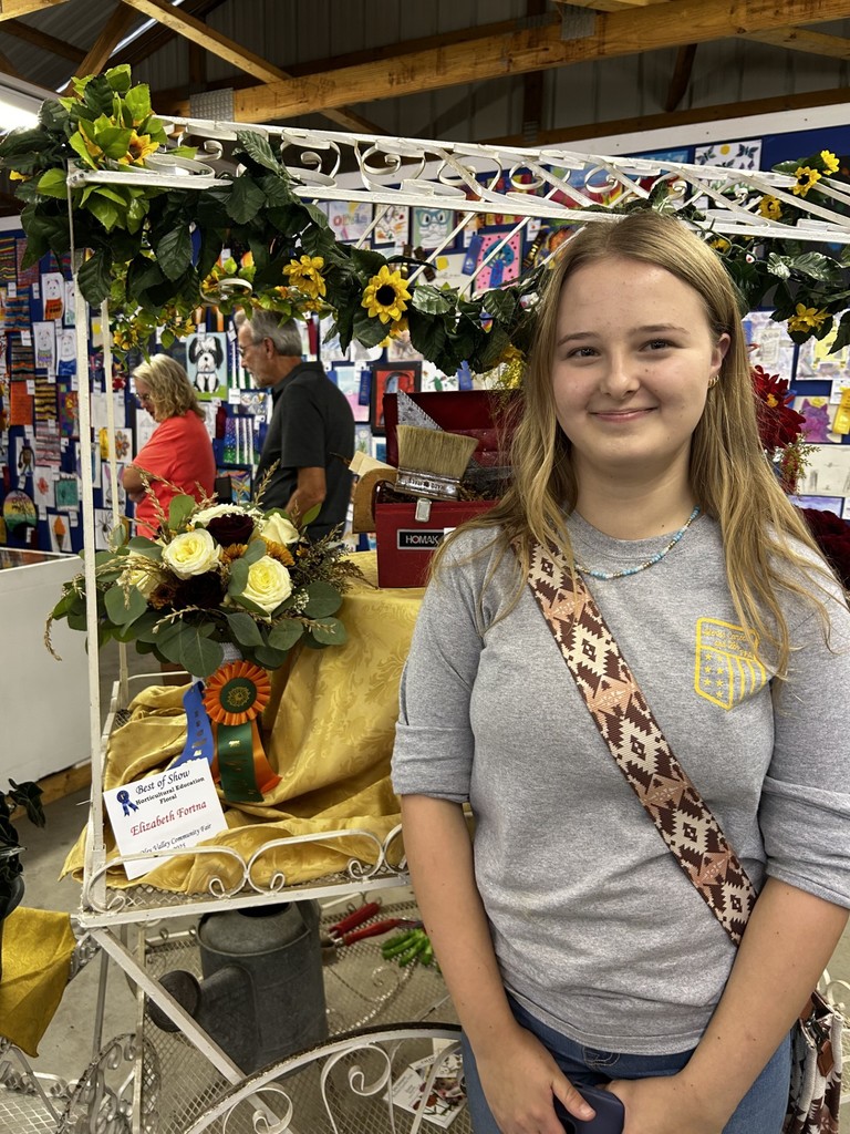 A smiling young woman, Elizabeth Fortna, stands next to her award-winning floral arrangement of roses and greenery on gold fabric. Her arrangement is decorated with a Best of Show rosette and a blue first-place ribbon at the Oley Valley Community Fair.