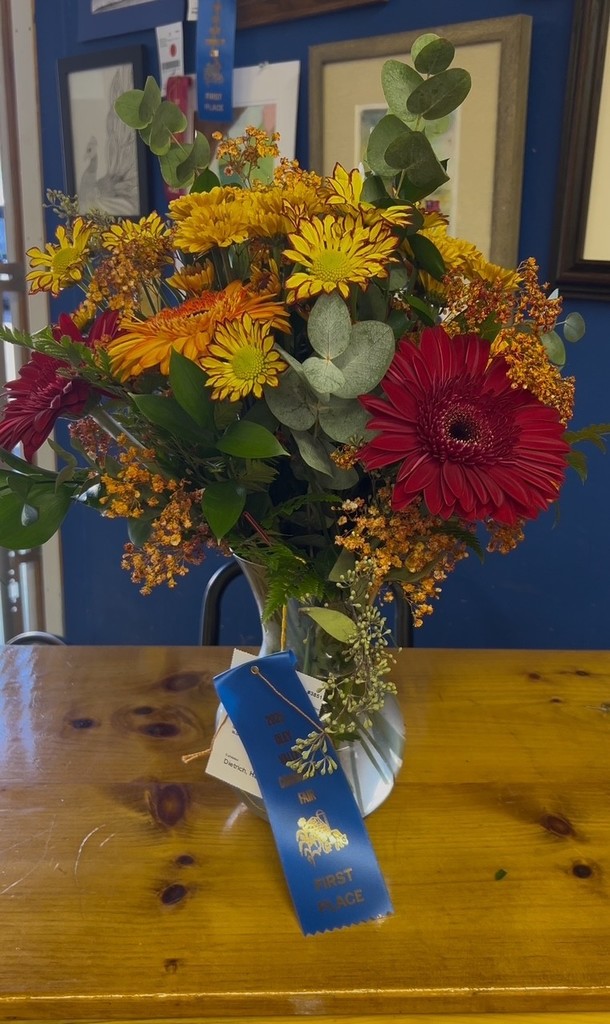 A tall arrangement of yellow, orange, and red flowers with greenery and eucalyptus displayed in a white pitcher vase. A blue first-place ribbon from the 2025 Oley Valley Community Fair is attached with exhibitor tag reading “Hannah Dietrich.”