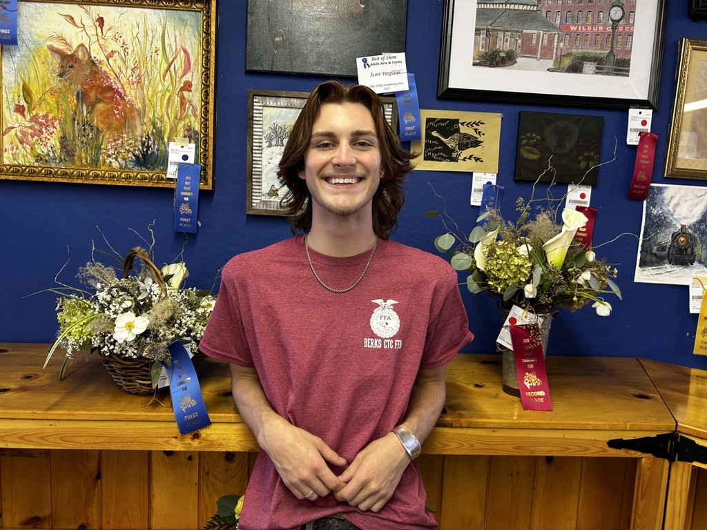 A young man, Logan Jones, stands smiling in front of two floral arrangements. One basket arrangement behind him has a blue first-place ribbon, and another vase arrangement has a red second-place ribbon.