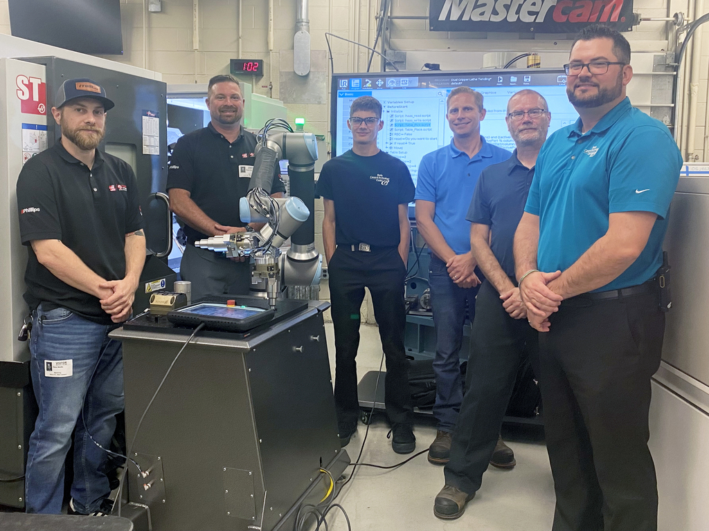 Six men stand in a machining lab next to a gray UR10e collaborative robot on a black base. Behind them are CNC machines, a large screen with programming software, and a Mastercam sign on the wall. The group includes instructors, industry partners, and a student, all smiling at the camera.