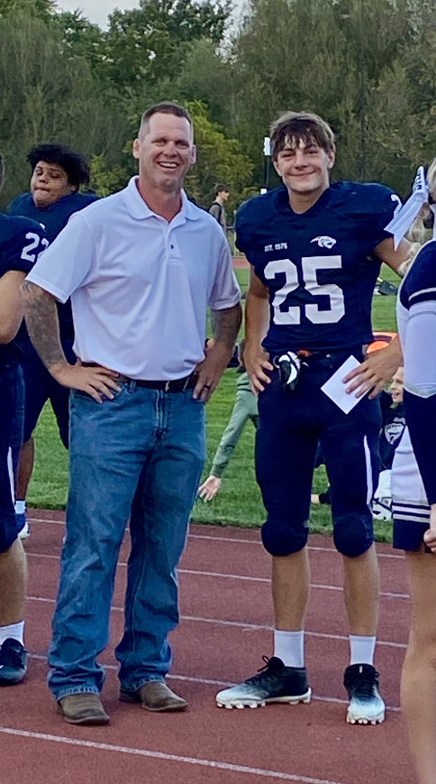 A high school football player in a navy blue uniform, number 25, stands on a track smiling with his arm at his side. Next to him is an adult man in a white polo shirt and jeans, also smiling with his hands on his hips. They are outdoors on the sideline of a football field, with other players and cheerleaders visible in the background.