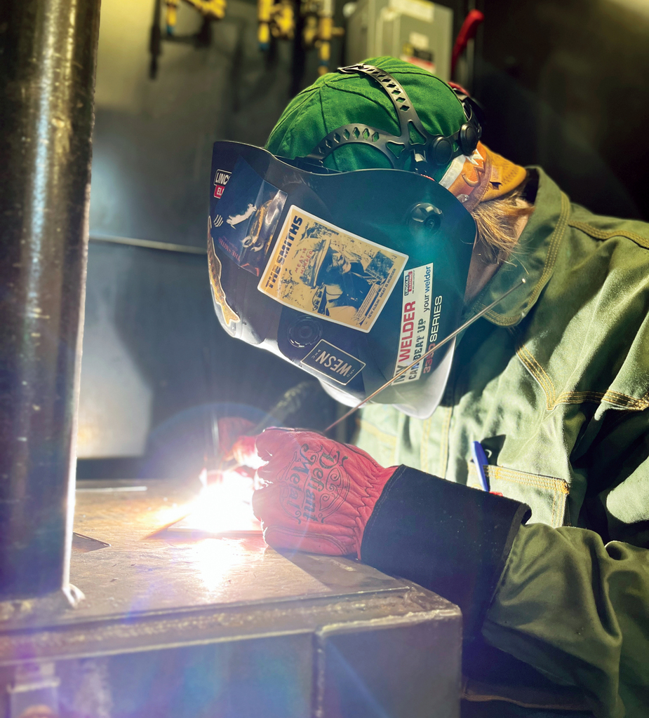 A welding student wearing protective gear, including a green cap, face shield with stickers, and red safety gloves, works on a piece of metal in a welding booth. Bright sparks and light from the welding torch illuminate the workspace.