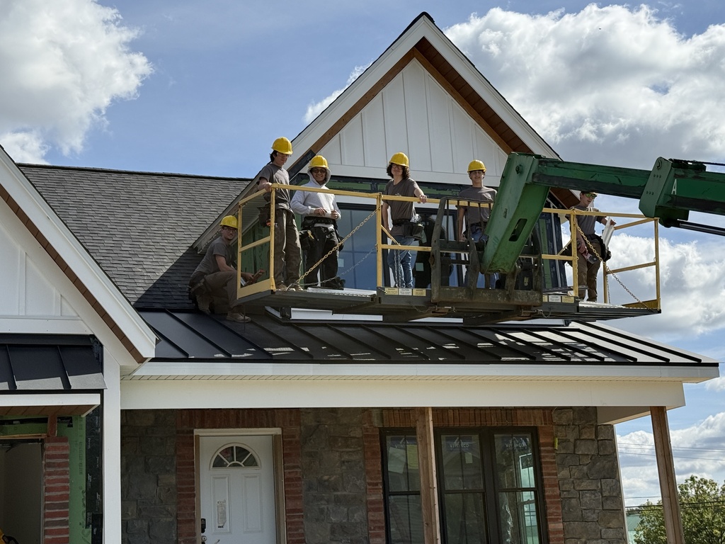 Close-up of several Building Construction Occupations students standing on a raised work platform. They are wearing yellow hard hats and safety gear as they prepare to apply siding to the upper portion of the house. The students smile and look toward the camera.
