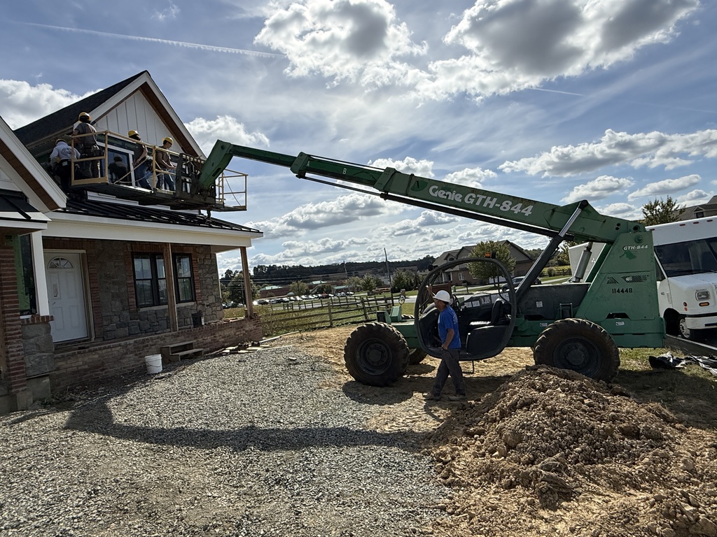 A green forklift extends a platform holding students in hard hats as they work on the siding of the upper story of the house. A worker in a blue shirt and white hard hat stands near the machine. The sky is filled with scattered clouds, and construction equipment and materials surround the site.