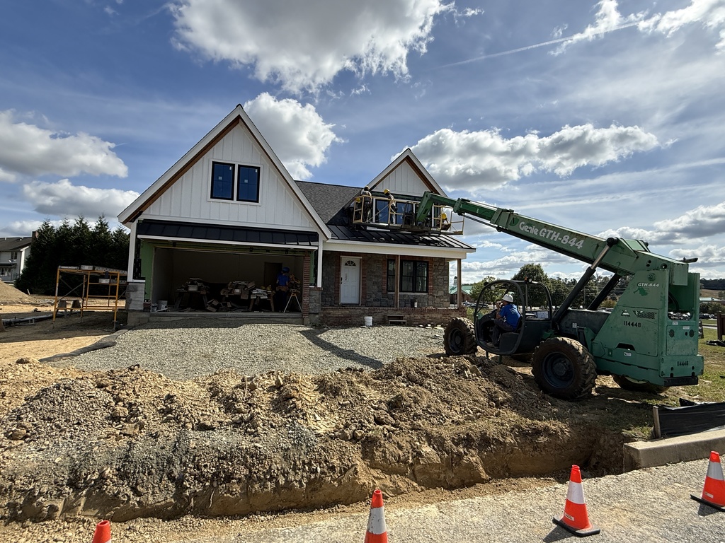 A wide shot of a two-story house under construction. The home has white siding, stone accents, and black trim. A green telescopic forklift is lifting a work platform with students in hard hats as they install siding on the upper level. Construction materials are scattered in the garage, and traffic cones mark the work area.