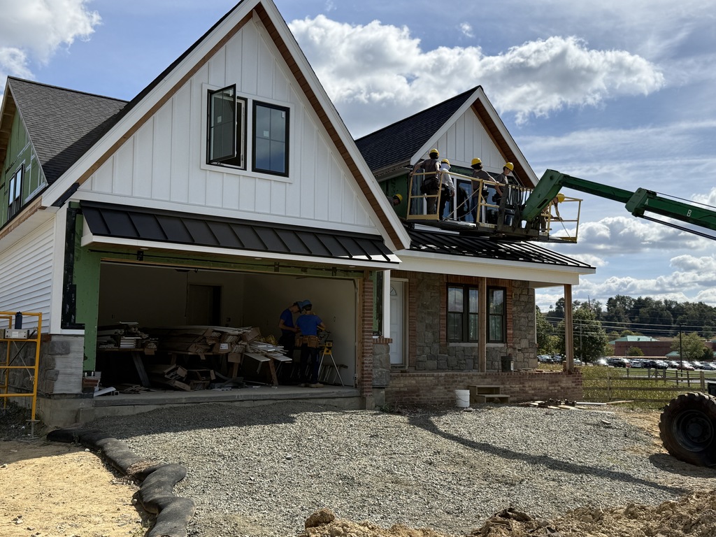 Mid-range view of the house with students working on the upper level from a raised platform. Two students are also working inside the garage with construction tools. White siding is partially installed along one side of the home.