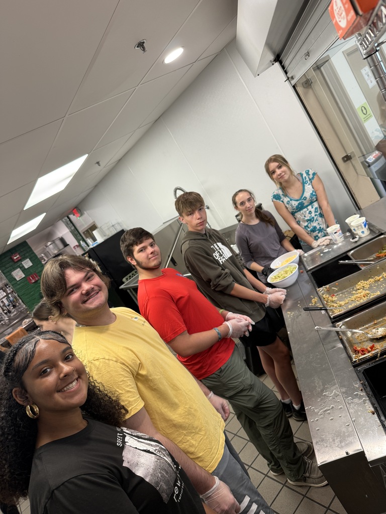 Students line up behind serving trays at a counter, smiling as they prepare to serve the meal they cooked.