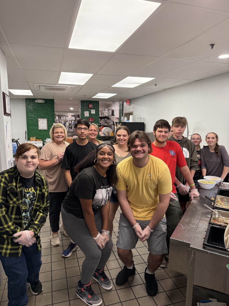 A larger group of students and staff gather in the kitchen for a cheerful group photo, some crouching and others standing, surrounded by cooking equipment and trays of food.