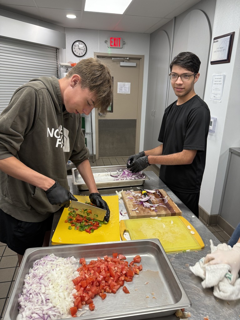 Two students work together slicing onions and peppers on yellow cutting boards, focused on meal prep in a kitchen environment.