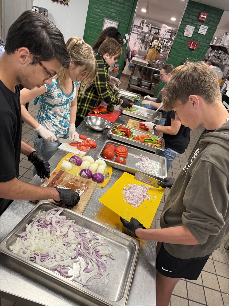 Several students line up at prep tables chopping tomatoes, onions, and peppers. The workspace is organized with colorful cutting boards and trays of fresh ingredients.