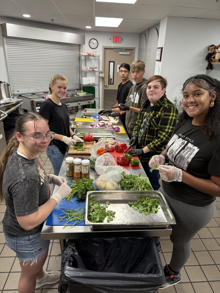 A group of students wearing gloves stand side-by-side at a stainless-steel prep table, smiling as they chop vegetables like peppers, onions, and cilantro in a commercial kitchen.