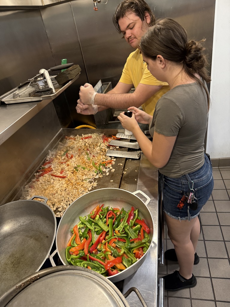 Two students stand over a flat-top grill sautéing chicken and peppers, with large pots of sliced vegetables beside them.
