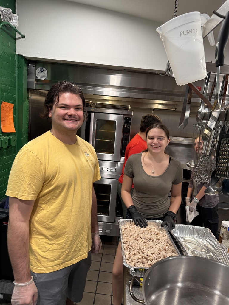 Two students stand behind a large tray of cooked chicken in a commercial kitchen, smiling as they prepare food near an oven.