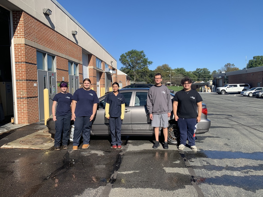 Five students stand in a row outside the BCTC automotive shop next to a gray car on a sunny day, smiling for the camera.