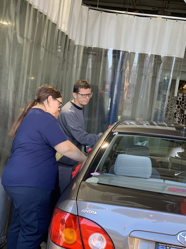 Two students wearing safety glasses and BCTC uniforms work together to wipe down the rear side of a gray Toyota sedan inside an automotive bay.