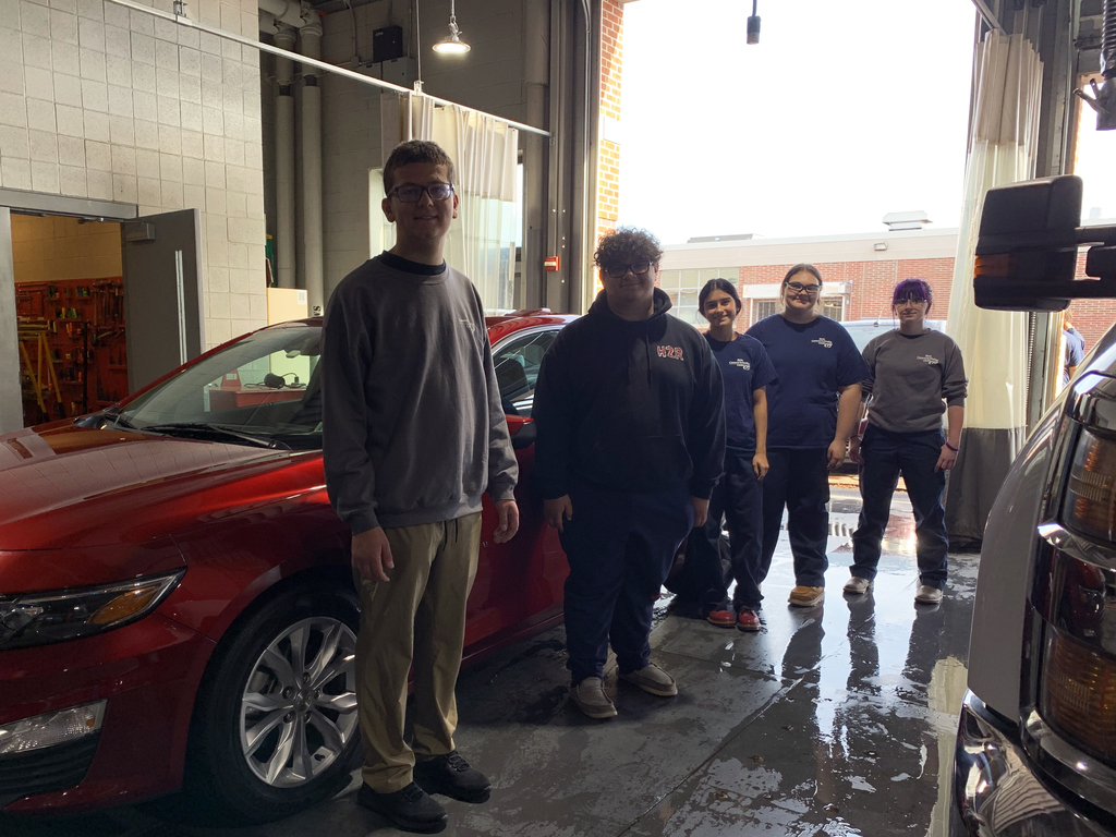 A group of five students stands inside the BCTC automotive bay beside a red car, smiling after completing a detailing project.