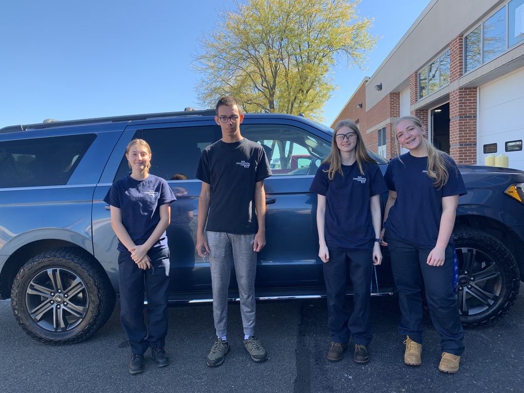 Four students wearing BCTC shirts stand in front of a blue SUV outside the automotive shop, posing together under clear blue skies.