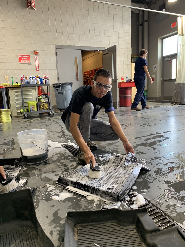 A student wearing safety glasses kneels on the shop floor scrubbing a black rubber car mat with a brush in a bucket of soapy water.