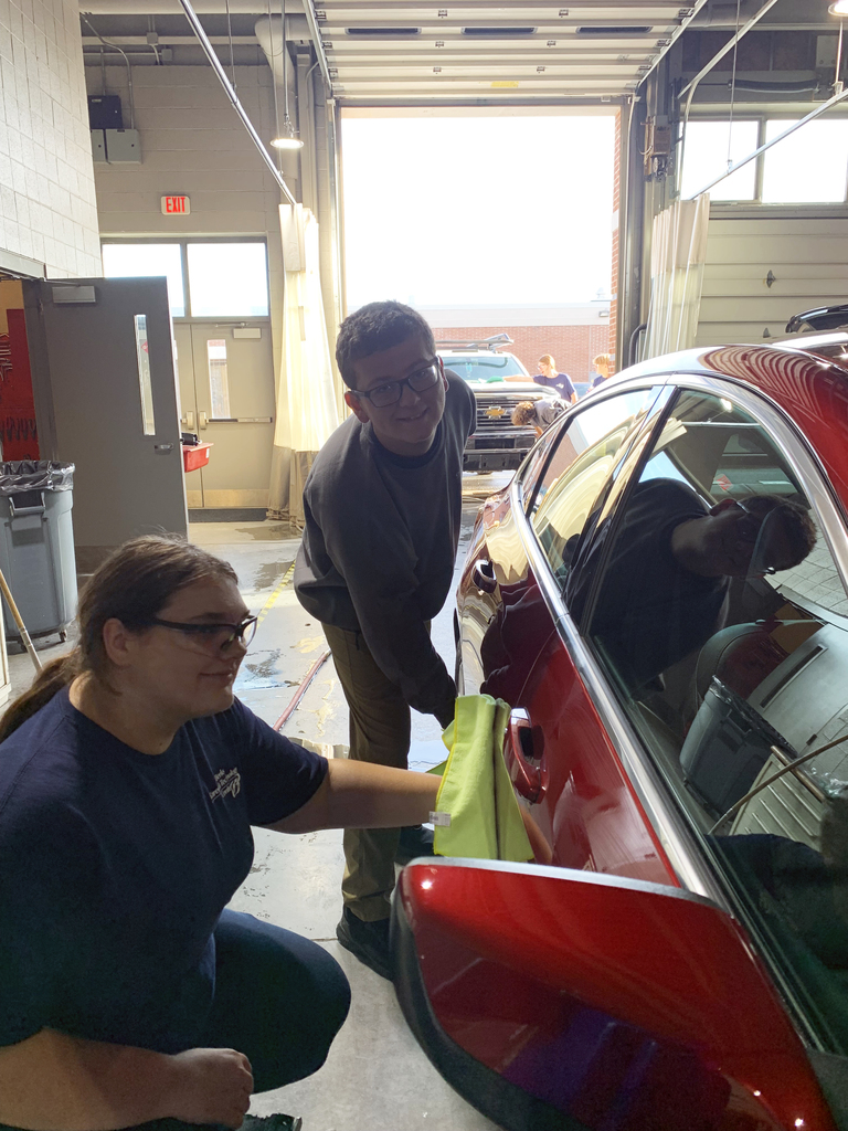 Two students wearing BCTC uniforms wipe the side of a shiny red car with a bright yellow microfiber cloth inside the automotive shop.