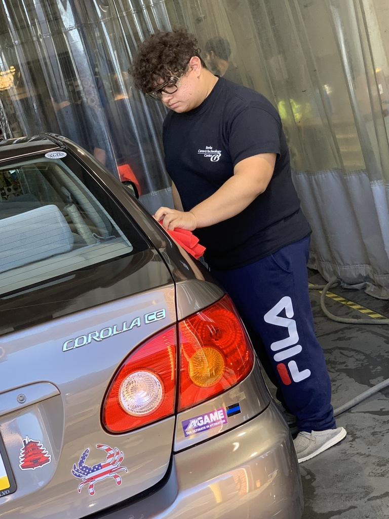 A student in a dark BCTC T-shirt and blue FILA pants uses a red cloth to clean the rear corner of a gray Toyota Corolla inside the shop.