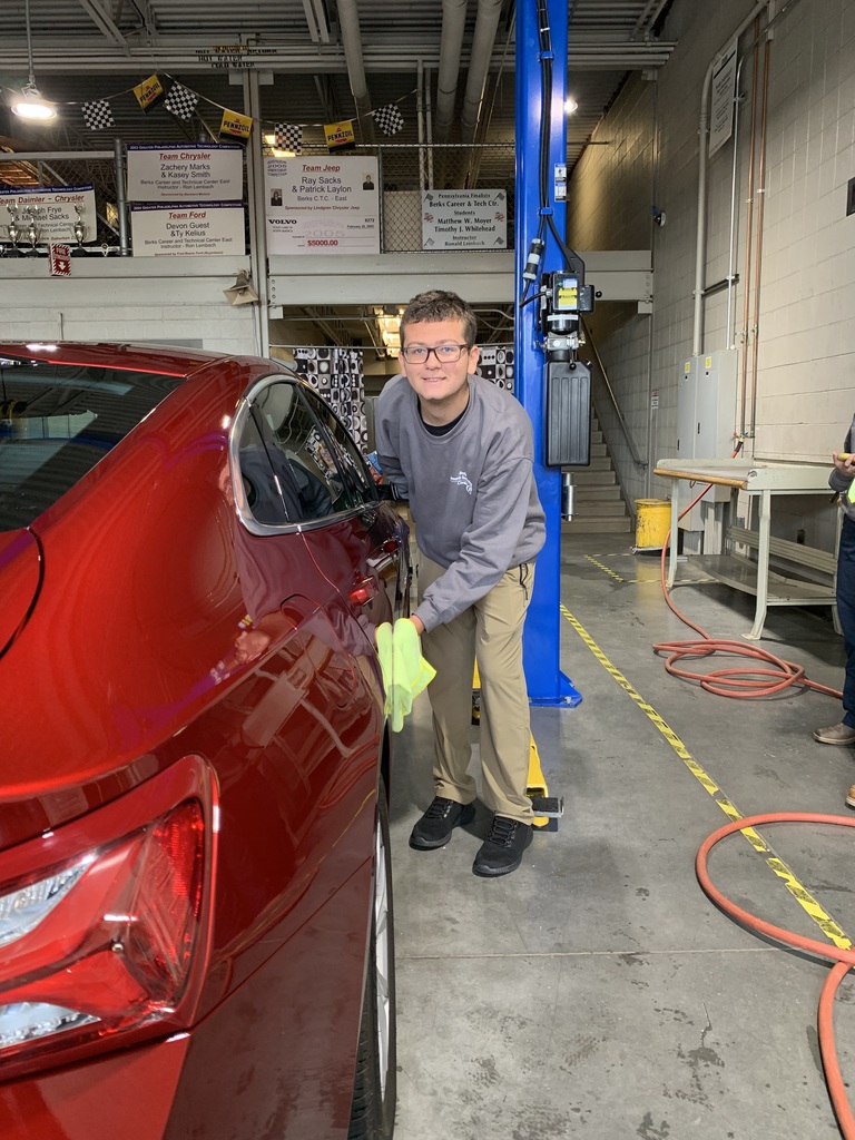 A student wearing safety glasses and a gray BCTC sweatshirt smiles while drying the side of a red car with a yellow cloth inside the automotive bay.