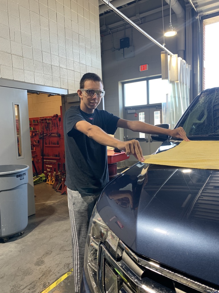 A student wearing safety glasses and a black shirt uses a yellow towel to wipe down the hood of a blue SUV inside the BCTC automotive bay.
