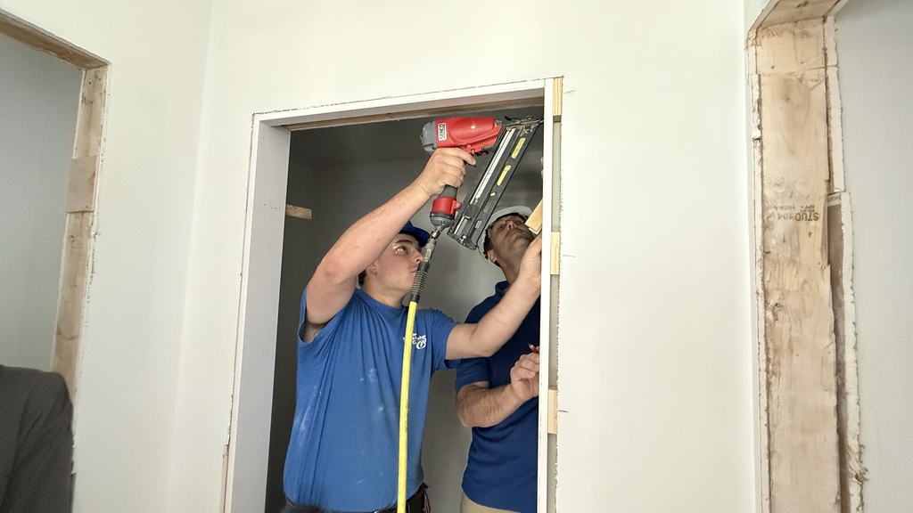 A Carpentry student in a blue shirt uses a nail gun to secure a door jamb while another person observes and assists. The door frame shows exposed wood as part of the installation process.