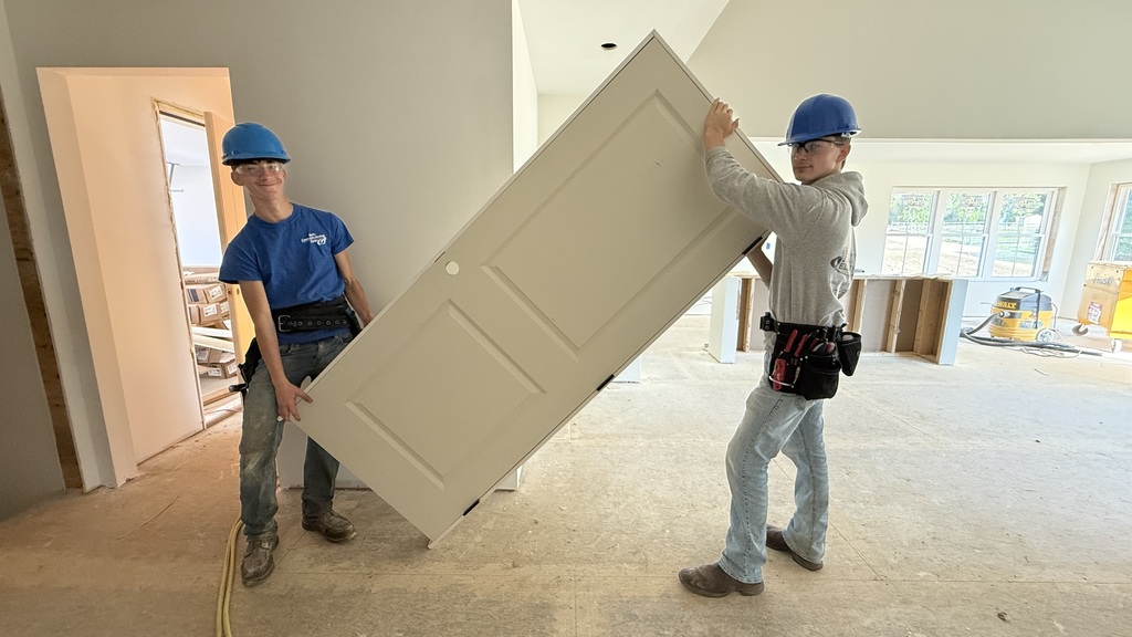 Two Carpentry students wearing blue hard hats and safety glasses carry a white door inside the Student-Built House. The unfinished interior shows exposed flooring and construction tools in the background.