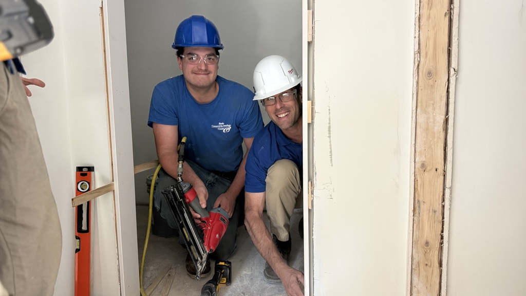 A Carpentry student and instructor wearing hard hats crouch in a doorway, smiling toward the camera while working with a nail gun and drill. Construction materials and tools are visible around them.