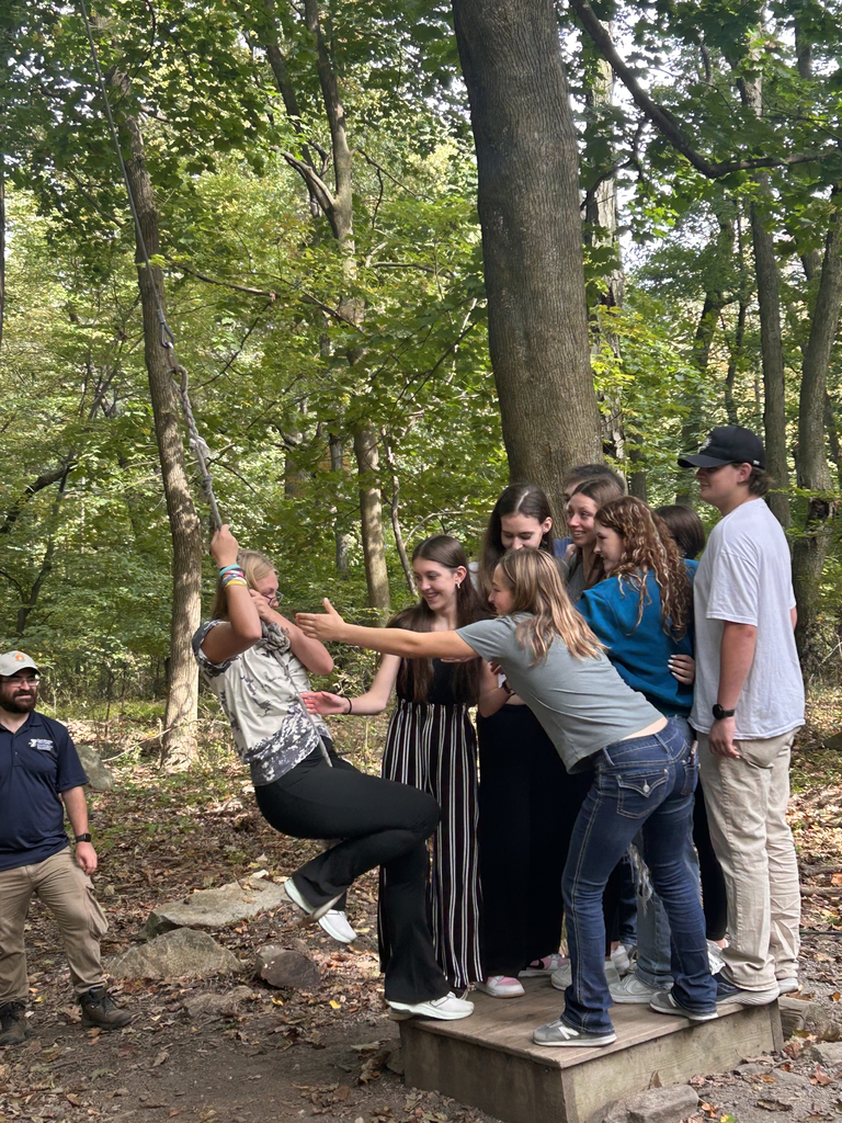 A group of NTHS students assist a classmate swinging on a rope during a trust-building challenge. Several students reach out to catch her while an instructor observes nearby.