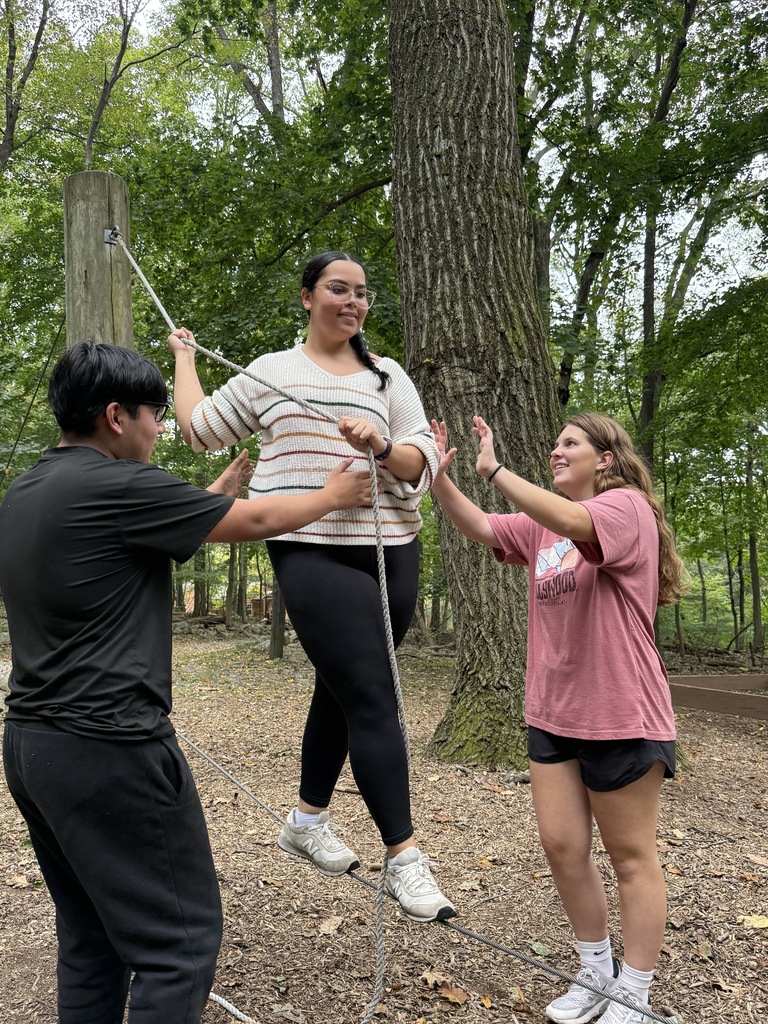 An NTHS student balances carefully on a low rope between two trees while classmates support and guide her from both sides.