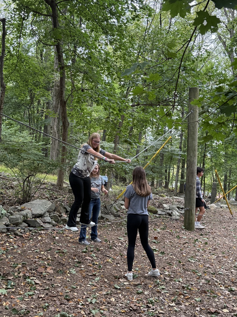 An NTHS student walks across a tightrope between trees while classmates offer verbal and physical support nearby in a wooded setting.