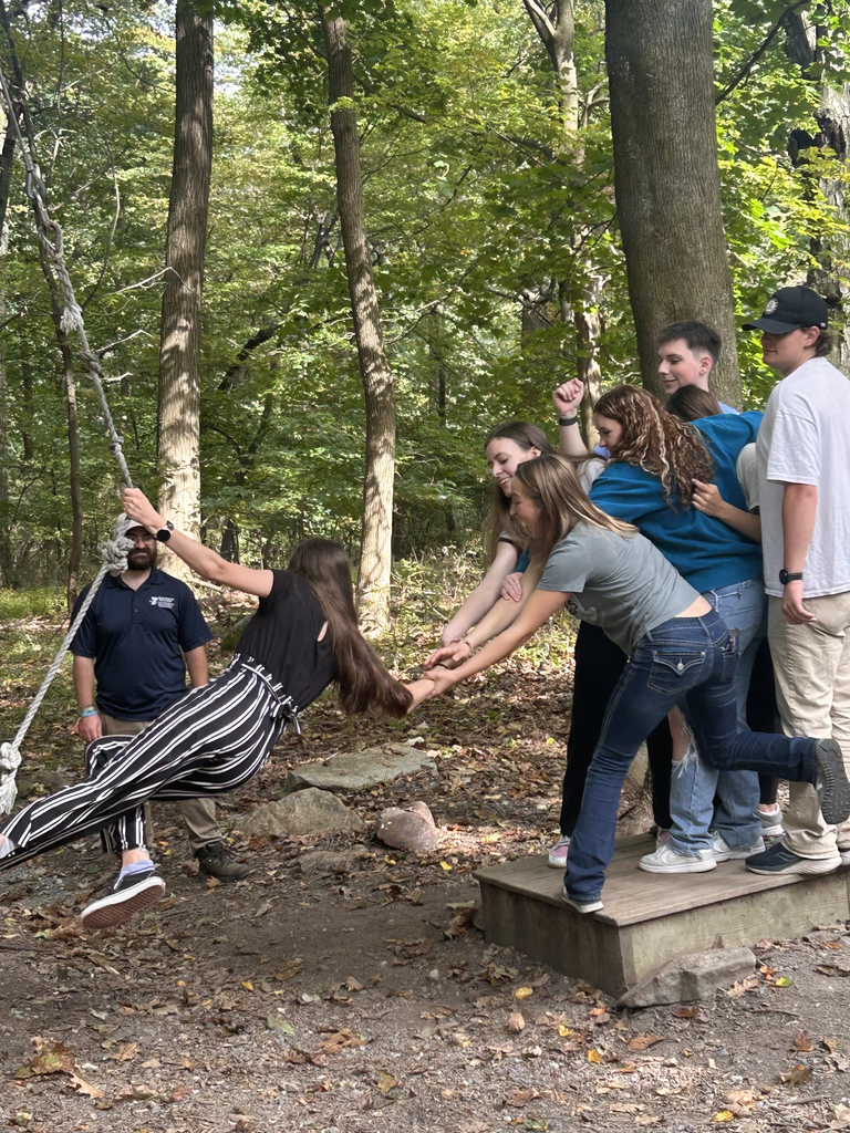 A group of NTHS students participate in a rope swing activity in the woods. One student swings forward while teammates extend their arms to catch her safely.