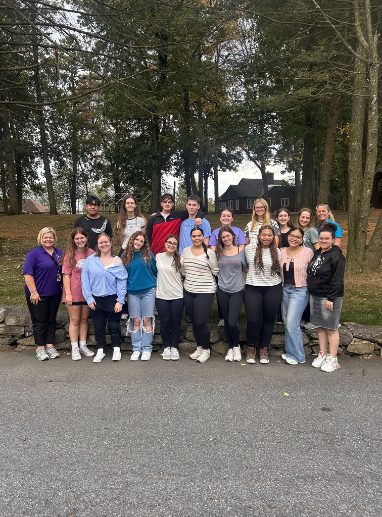 A large group of National Technical Honor Society students and advisors stand together outdoors in front of a stone wall at South Mountain YMCA. The group smiles for a photo surrounded by tall trees and rustic cabins in the background.