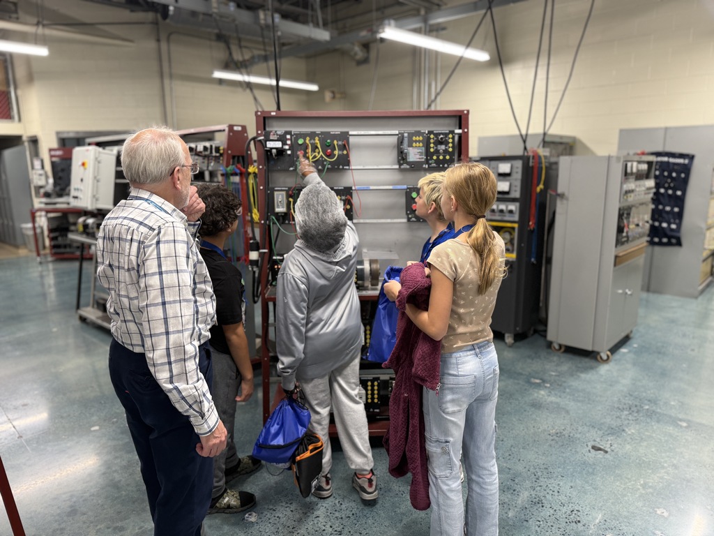 Students explore a panel of electrical components in BCTC’s Mechatronics Engineering Technology lab. An instructor observes as one student points at illuminated buttons and wiring, while others look on with curiosity.