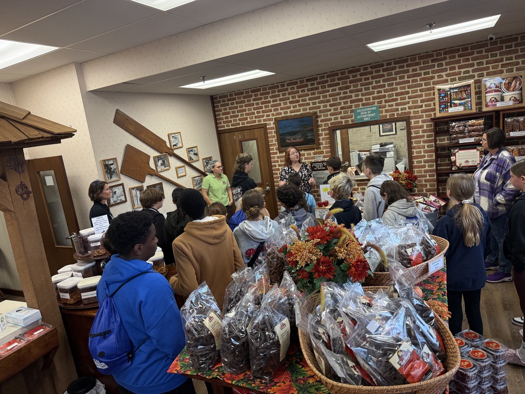 A group of students stands inside the Tom Sturgis Pretzels retail shop, surrounded by baskets of packaged pretzels. They are listening to a staff member describe the company’s history and production process.