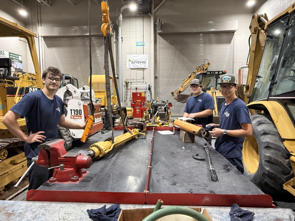 Three Heavy Equipment Technology students stand at a long red workbench surrounded by heavy machinery, working together to remove and repair hydraulic cylinders. All are wearing safety glasses and navy BCTC shirts.