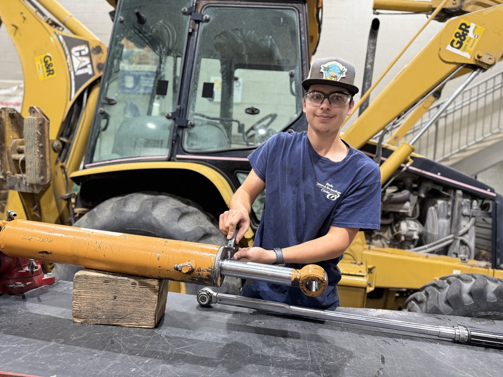 A Heavy Equipment Technology student wearing safety glasses and a dark blue BCTC shirt uses a wrench to remove a hydraulic cylinder on a workbench. A yellow backhoe is parked behind him inside the shop.