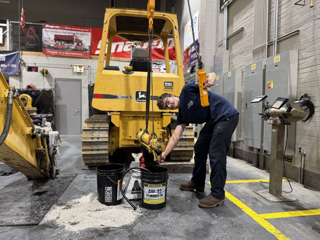 A Heavy Equipment Technology student operates a hoist to lift a hydraulic cylinder near a yellow John Deere crawler. Buckets of hydraulic oil and absorbent material are placed on the shop floor beneath the equipment.