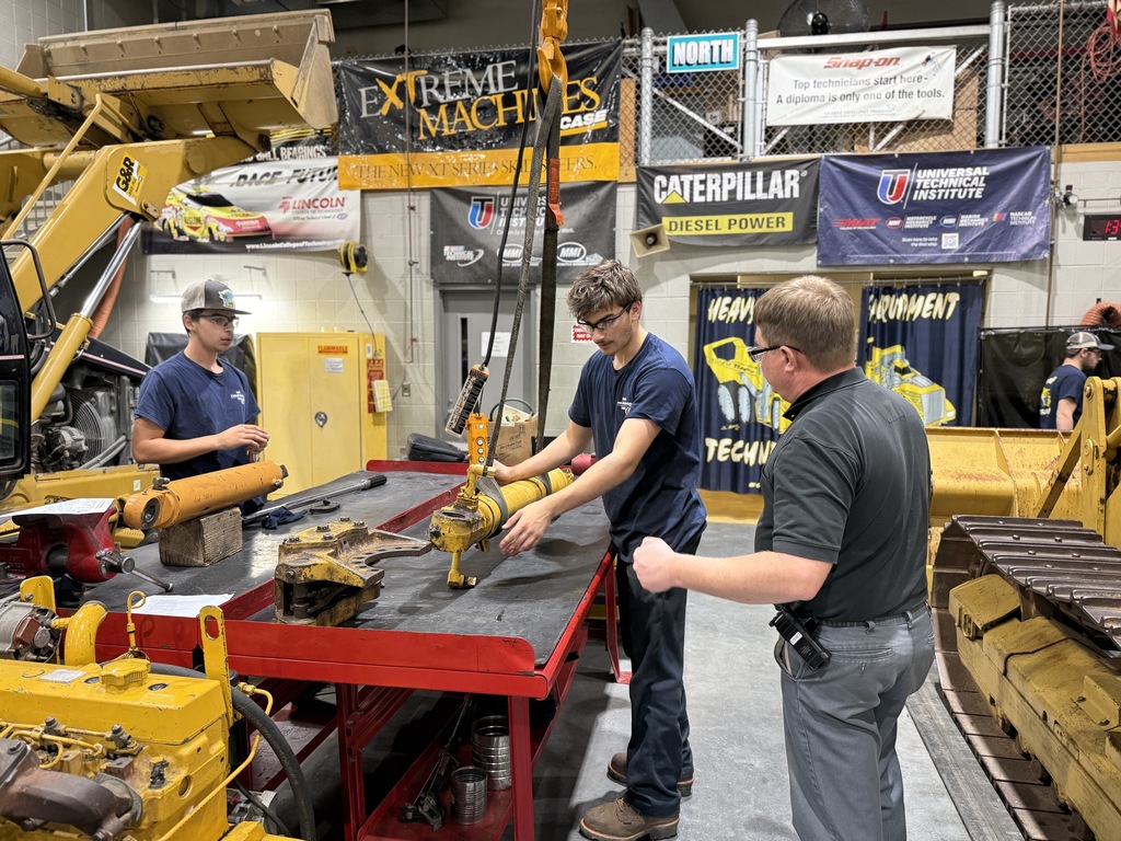 Two Heavy Equipment Technology students work together on a hydraulic cylinder at a red workbench while an instructor observes and provides guidance. Large yellow equipment and training banners hang in the background of the shop.